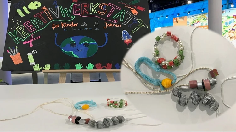 Auf dem Bild liegen gebstaltete Papierperlenketten auf dem Tisch und im Hintergrund steht eine Tafel.